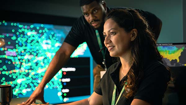Female employee typing and male employee standing behind her looking at the computer in the command center